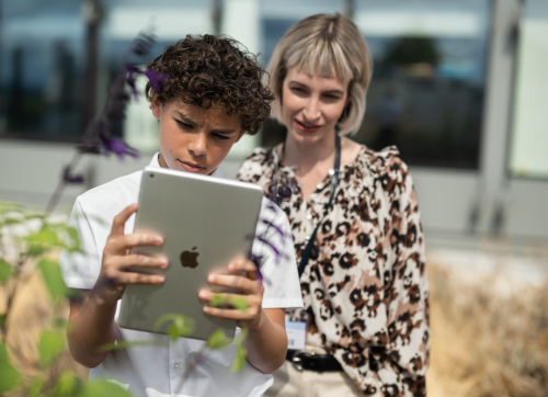 A Student Using Ipad Outside The Schooll For Project Work With His Teacher Standing Behind Him
