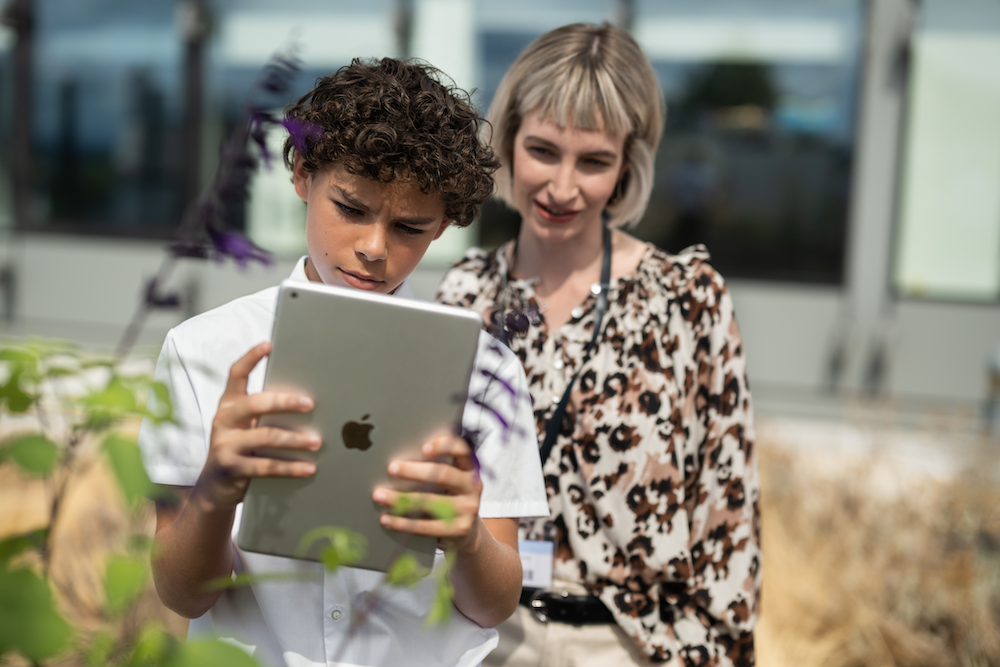 A student using ipad outside the schooll for project work with his teacher standing behind him