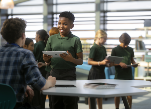 A Student Smiling Talking To Their Teacher At His Desk While Holding An Ipad