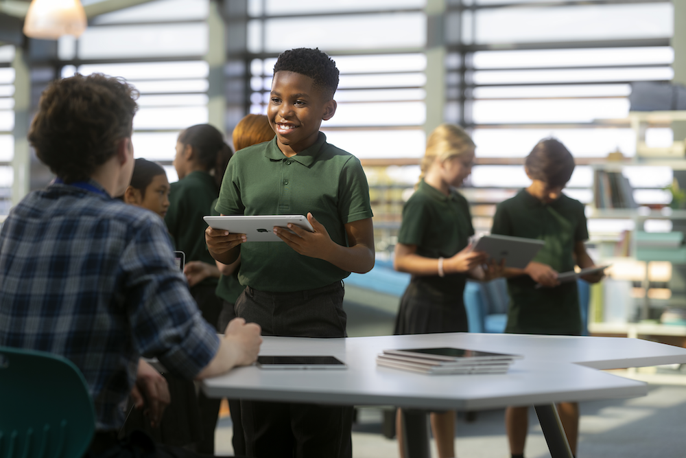 A student smiling talking to their teacher at his desk while holding an ipad